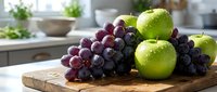 Fresh green apples and dark red grapes with water drops on a wooden cutting board in a bright modern kitchen.
