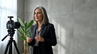 Middle-aged woman with long gray hair and Caucasian ethnicity wearing black blazer, speaking or presenting in front of video camera on tripod in modern studio with concrete wall and green plant.