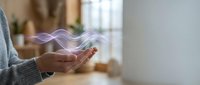 Woman hands holding smartphone with purple digital wave energy flowing above device in modern home interior for technology and communication concepts.