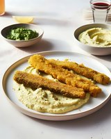 Crispy golden breaded fish sticks served on creamy hummus with side bowls of green herb sauce and additional hummus on white table setting.