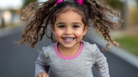 Young girl with curly hair in pigtails wearing gray and pink shirt smiling outdoors on blurred street background, showing natural joy and happiness in childhood moment.