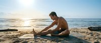 Young Caucasian man practicing yoga stretching pose on sandy beach at sunrise, ocean waves in background, healthy lifestyle concept.