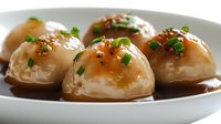 Steamed dumplings with glossy brown sauce and fresh herbs in white bowl. Traditional Asian cuisine ready for restaurant menu or food blog content.