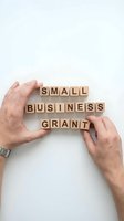 Hands protecting wooden letter blocks spelling small business grant on white background for entrepreneurship and funding concepts.