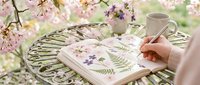 Young woman with fair complexion painting watercolor flowers in sketchbook outdoors among blooming cherry blossoms, spring botanical art and nature inspiration for creative projects.