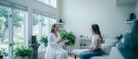 Two women having friendly conversation in bright modern living room with large windows and green plants creating comfortable home atmosphere.