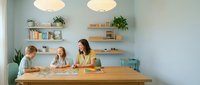 Mother and two children doing homework together at wooden table in modern mint green kitchen with floating shelves and pendant lights.