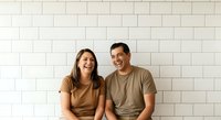 A young couple, Caucasian male and female in their twenties, wearing casual brown and tan t-shirts, smiling together against a white subway tile wall. Suitable for lifestyle, relationship, and casual apparel marketing.