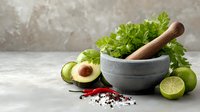 Fresh ingredients for guacamole preparation with stone mortar, avocado, lime, cilantro, and spices on concrete background.