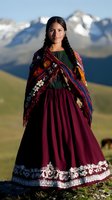 Young Latin American woman in traditional Andean dress with colorful woven shawl and burgundy skirt standing against snow-capped mountain backdrop in natural lighting.