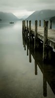 Weathered wooden pier extending into calm alpine lake surrounded by misty mountains and distant boat, moody landscape for travel and nature projects.