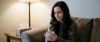 Young woman with long dark hair and South Asian ethnicity sitting on beige sofa, holding tissue box, appearing unwell or emotional in warm home interior setting.