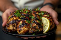 Grilled chicken thighs with charred marks, garnished with fresh cilantro and lime wedges served on black plate, hands holding dish in dark moody setting.