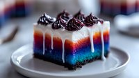 Colorful layered dessert bar with chocolate frosting swirls and white drizzle on white plate. Vibrant blue, red and white gradient creates striking visual effect.
