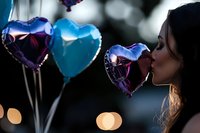 Young woman kissing heart shaped balloon at twilight with bokeh lights background, romantic mood with blue and purple foil balloons creating dreamy atmosphere.