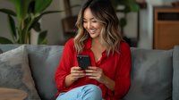 Young Asian woman in red blouse sitting on gray couch, smiling while using smartphone. Natural indoor lighting and home environment creates relaxed lifestyle scene.