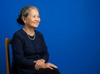 Mature woman with gray hair and Asian ethnicity, wearing dark navy cardigan and pearl necklace, seated against solid blue background. Professional portrait for corporate, business, and editorial use.