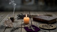 Mystical altar setup with burning candle, tarot cards, incense smoke, crystals and occult symbols on rustic wooden table for spiritual divination practices.