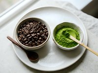Two ceramic bowls on white plate showcase roasted coffee beans and vibrant green matcha powder with wooden spoon, representing contrasting beverage choices.