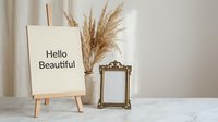 Wooden easel with Hello Beautiful text, pampas grass in vase, and golden frame on marble surface, warm neutral boho interior decor.