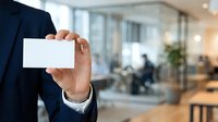 Professional man in dark suit holding blank white business card in modern office environment with blurred colleagues and glass partitions in background for corporate branding and networking.
