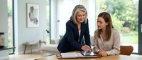 Mature woman with long gray hair and younger woman with brown hair reviewing documents together at wooden table in bright modern office space with large windows.