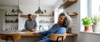 African American woman relaxing at kitchen counter while partner cooks in modern home interior with natural lighting and wooden accents.