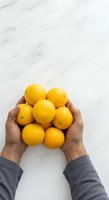 Person holding fresh yellow lemons against marble background. Overhead view showing hands in gray sleeves cradling ripe citrus fruits for healthy eating, cooking, and beverage concepts.