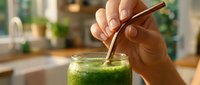 Person with medium skin tone stirring fresh green smoothie or juice with wooden spoon in bright kitchen setting. Healthy beverage preparation and wellness lifestyle concept.