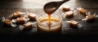 Homemade salted caramel sauce dripping from wooden spoon into glass jar surrounded by golden caramel candies on dark rustic surface for dessert recipes.