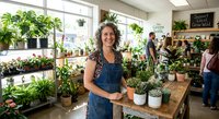 Young Caucasian woman florist in denim apron smiling while working with potted plants in bright greenhouse nursery shop interior with customers browsing.