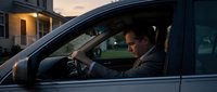 Professional businessman driving luxury car during evening commute in residential area with warm house lights creating atmospheric urban lifestyle scene.