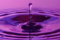 Single water drop impact creating crown splash and column on purple liquid surface with concentric ripple rings in macro photography. Frozen motion capture.