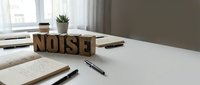 Wooden letter blocks spelling NOISE on white desk with notebooks, pens and plant. Office workspace concept for sound pollution, distraction and productivity challenges.