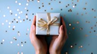Female hands holding small wrapped present with golden bow on blue background with festive confetti and bokeh lights. Celebration concept.