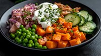 Fresh bowl with roasted sweet potato cubes, green peas, cucumber slices, red onion, and sour cream garnished with herbs on black plate for healthy lunch.