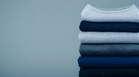 Stack of neatly folded casual cotton sweatshirts and t-shirts in various shades of blue and gray against neutral background, minimalist fashion composition.