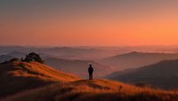 Lone hiker silhouette standing on golden hillside at sunset with layered mountain ranges stretching into distance under warm orange sky.