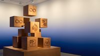 Wooden blocks with sustainability symbols stacked in pyramid formation on display platform against gradient blue wall in modern gallery space.