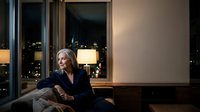 Professional blonde businesswoman in dark suit relaxing on sofa in modern office with city lights view through windows during evening hours.