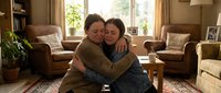 Two women embracing warmly in cozy living room with natural light, showing love and connection between family members or close friends in comfortable home setting.
