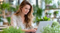 Young Caucasian woman with long wavy hair working on laptop in greenhouse surrounded by lush green plants, creating peaceful home office environment.