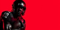 African American football player in protective gear against vibrant red background, dramatic lighting creating powerful sports portrait.