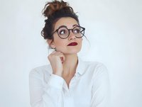 Young professional woman with glasses and messy bun looking up thoughtfully while touching chin, wearing white shirt against neutral background for business concepts.