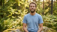 Caucasian man with blonde beard wearing light blue t-shirt sitting on rock in forest with green trees and natural sunlight, outdoor portrait for lifestyle and wellness content.