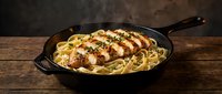 Grilled chicken breast sliced over creamy pasta in cast iron skillet on rustic wooden table for restaurant menu and cooking content.