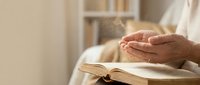 Elderly hands in prayer position over open book in peaceful home setting creating spiritual meditation moment for faith and devotion concepts.