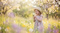 Young Caucasian girl in floral dress and straw hat blowing dandelion in sunny blooming garden with wildflowers in spring.