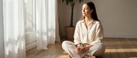 Young woman in beige pajamas meditating in lotus position on wooden floor by window with natural sunlight streaming through white curtains in modern bedroom.