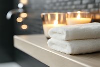 Folded white spa towels on wooden shelf with glowing candles creating relaxing atmosphere. Wellness and body care concept for spa treatments and massage.